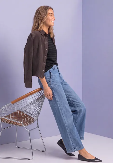 Frau in brauner Jacke, schwarzem Shirt und weiten Jeans lehnt lässig an einem Drahtstuhl vor lila Wand.