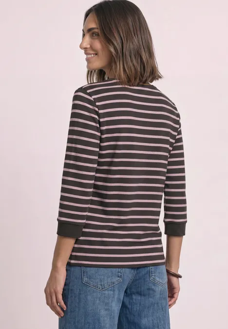Frau mit braunen Haaren von hinten, trägt gestreiftes Langarmshirt und Jeans, im Profil vor rosa Hintergrund.