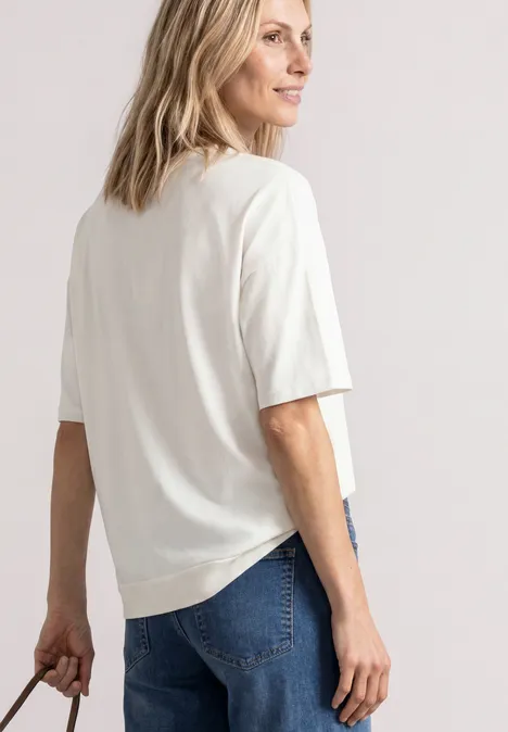 Vrouw opzij in wit T-shirt en spijkerbroek, met een tas in haar hand tegen een lichte achtergrond.