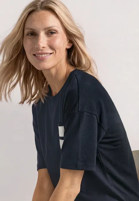 Blonde vrouw lacht in een donker T-shirt, zittend schuin en kijkend naar de camera.