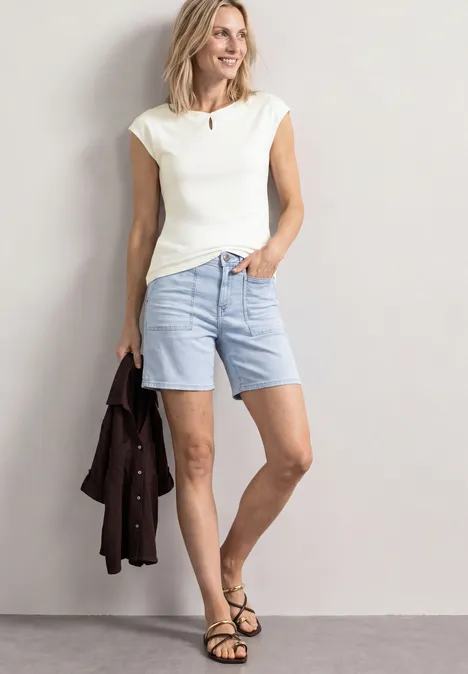 Femme en top blanc et short bleu clair, tient une veste marron, porte des sandales noires.