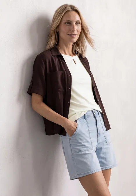 Personne en t-shirt blanc, chemise marron ouverte et short bleu clair appuyée contre un mur, mains dans les poches.