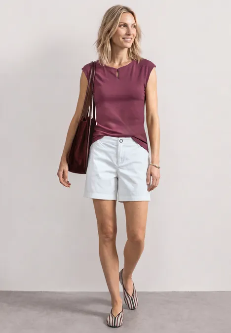 Shirt met afhangende schouders en uitsnijding dark clouded blush Vrouw met bordeaux top, witte short en slippers, draagt grote tas voor neutrale achtergrond.