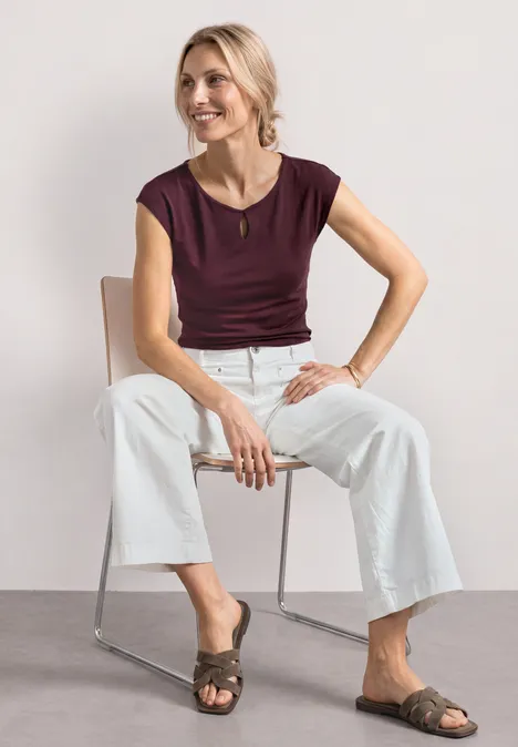 Shirt met afhangende schouders en uitsnijding rustic rouge Vrouw zit op metalen stoel, draagt een bordeaux top, witte wijdvallende broek en platte sandalen