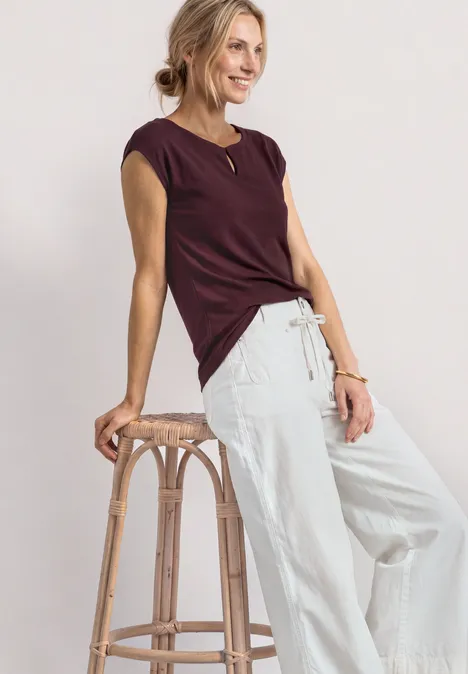 Shirt met afhangende schouders en uitsnijding rustic rouge Vrouw leunt op een houten kruk, draagt een bordeaux kort shirt en een lichte, wijde broek.