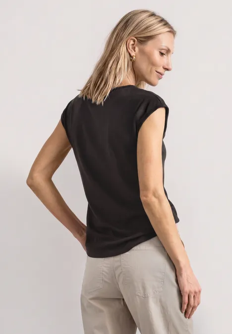 Shirt met afhangende schouders en uitsnijding black coffee Vrouw in zwart T-shirt en beige broek, van achteren gefotografeerd.