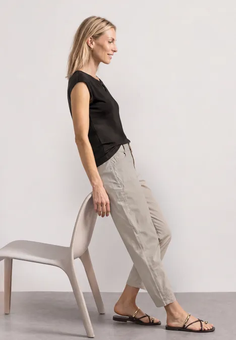 Shirt met afhangende schouders en uitsnijding black coffee Vrouw in zwart shirt en beige broek leunt zijwaarts tegen een stoel, draagt platte sandalen.