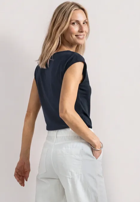 Une femme vue de dos porte un t-shirt noir et un pantalon blanc, mains dans les poches.