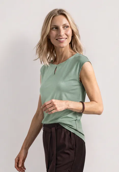 Femme en haut sans manches vert clair et pantalon foncé sur fond neutre.