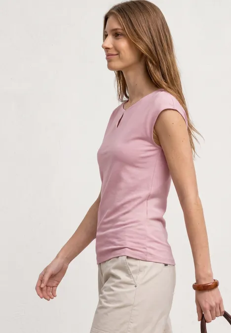 Profil latéral d’une femme en haut rose sans manches et pantalon beige devant un fond neutre.