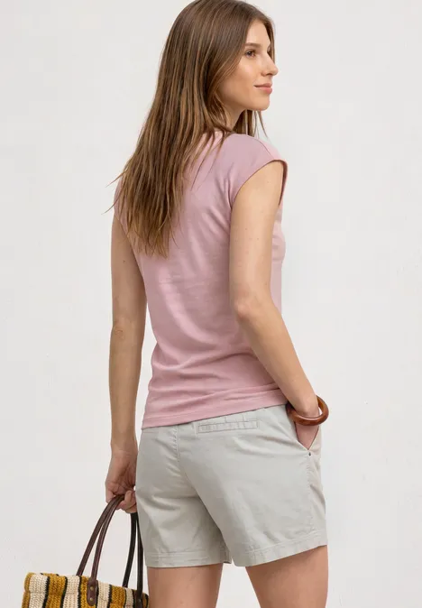 Femme de dos en t-shirt rose et short beige, tenant un sac à main marron devant un mur clair.