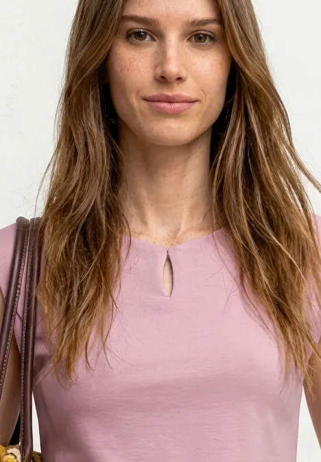 Femme en haut rose avec découpe à l'encolure, sac marron à l'épaule, cheveux longs bruns, fond blanc
