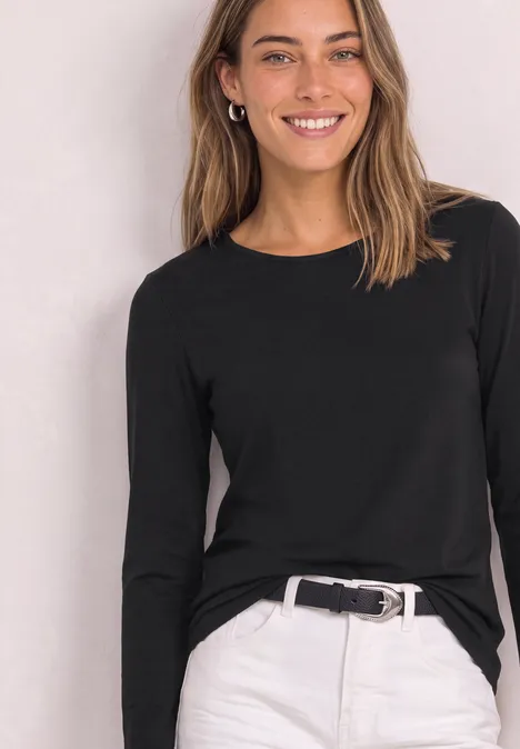 Eine Frau trägt ein schwarzes Langarmshirt und weiße Hose mit Gürtel und lächelt in die Kamera.