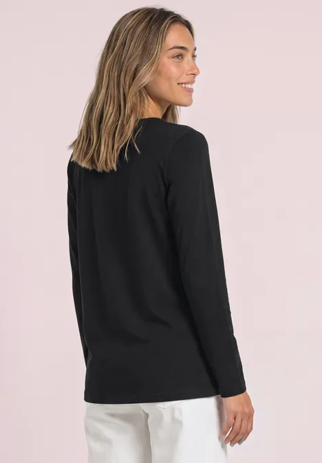 Frau in schwarzem Langarmshirt und weißer Hose, steht seitlich vor hellem, rosafarbenem Hintergrund.