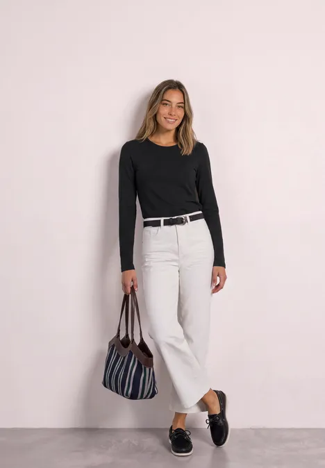 Frau in schwarzem Langarmshirt, weißer weiter Hose, schwarzem Gürtel, gestreifter Tasche und Loafers lächelnd.