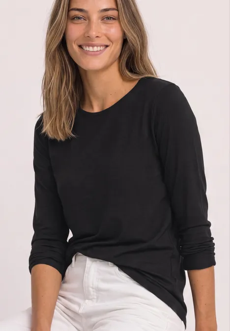 Eine lächelnde Frau in schwarzem Langarmshirt und weißer Hose vor hellem Hintergrund.