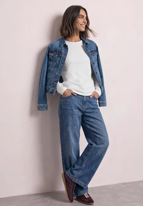 Frau in lässigem Denim-Look: weiße Bluse, Jeanshose und Jeansjacke, lächelnd an heller Wand lehnend.