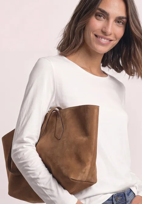 Frau in weißem Langarmshirt und Jeans trägt eine braune Wildleder-Tasche lässig unter dem Arm.