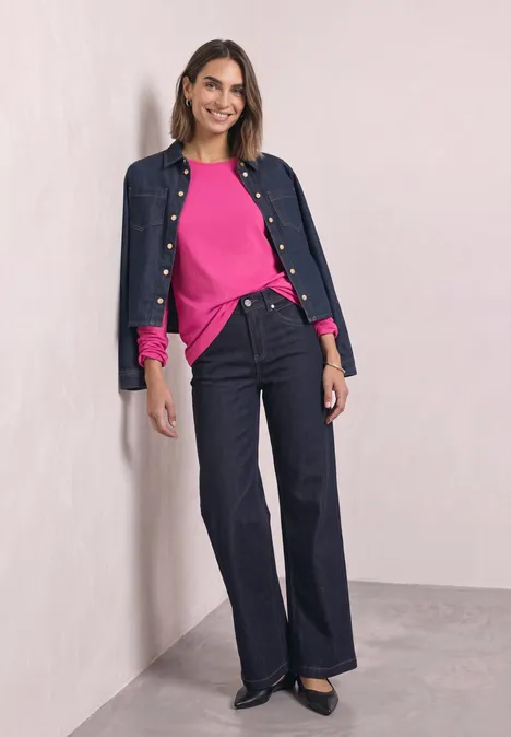 Basic Langarm Shirt in Unifarbe magenta dream Frau lehnt an Wand, trägt pinken Pullover, dunkle Jeans und offene Jeansjacke, lächelt.