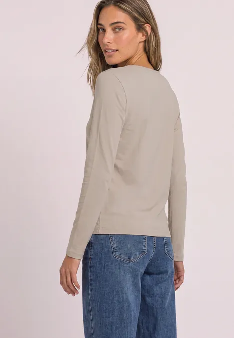Frau trägt beiges Langarmshirt und blaue Jeans, schaut über die Schulter vor hellem Hintergrund.