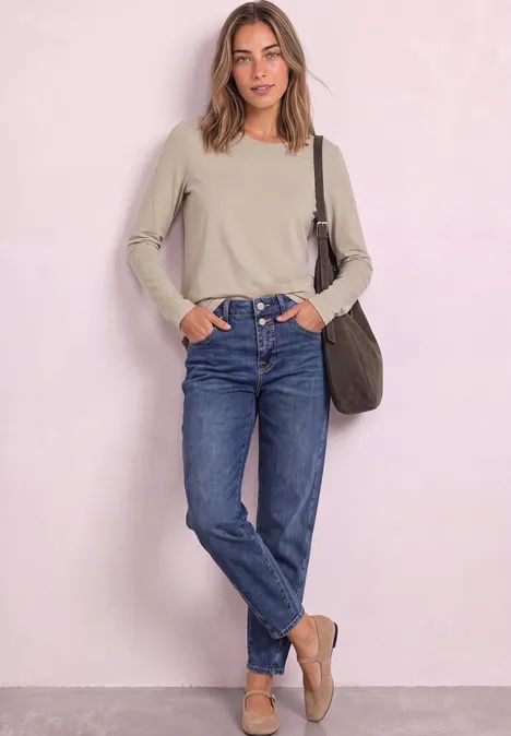 Junge Frau in beige Longsleeve, blauen Jeans und Loafers lehnt entspannt vor hellrosa Wand.