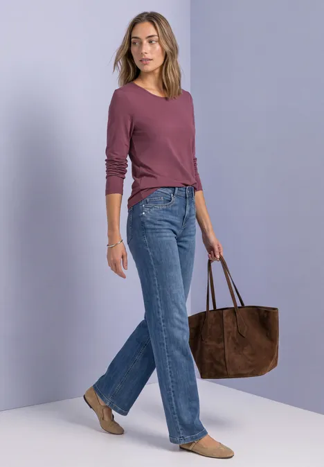 Frau in lila Oberteil, blauen Jeans und beigen Schuhen trägt eine große braune Wildledertasche.