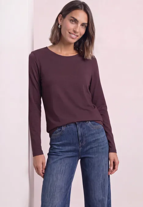 T-shirt basique à manches longues, couleur unie rustic rouge Femme portant un t-shirt manches longues bordeaux et un jean bleu, souriante, cheveux mi-longs et boucles d’oreilles.