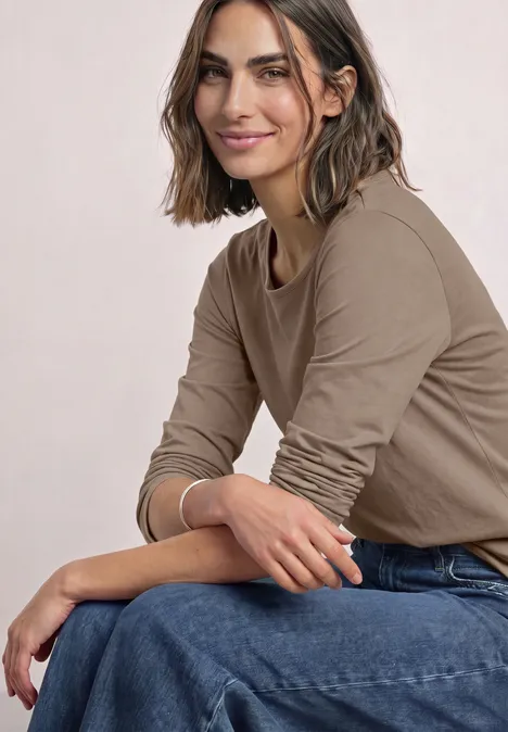 Basic Langarm Shirt in Unifarbe sanded amber Junge Frau mit schulterlangen braunen Haaren sitzt seitlich in blauer Jeans und beige Shirt, lächelt dezent.