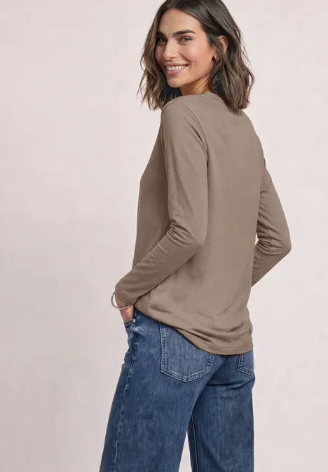Basic Langarm Shirt in Unifarbe sanded amber Eine lächelnde Frau in langärmligem beigem Oberteil und blauen Jeans blickt über die Schulter zur Kamera.