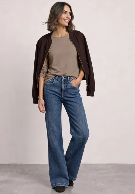 Basic Langarm Shirt in Unifarbe sanded amber Frau in weitem Jeans, olivgrünem T-Shirt und dunkelbrauner Jacke, lächelt selbstbewusst in neutralem Raum