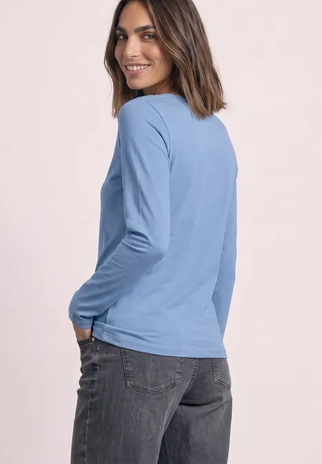 Frau im blauen Langarmshirt und dunklen Jeans steht seitlich vor rosa Hintergrund und lächelt.