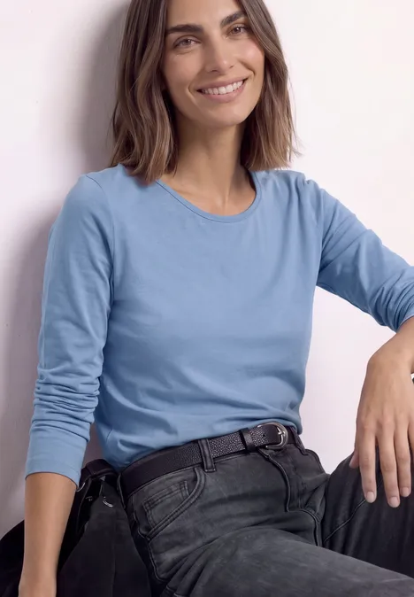 Frau lächelt in blauem Langarmshirt und dunkler Hose, lehnt entspannt.