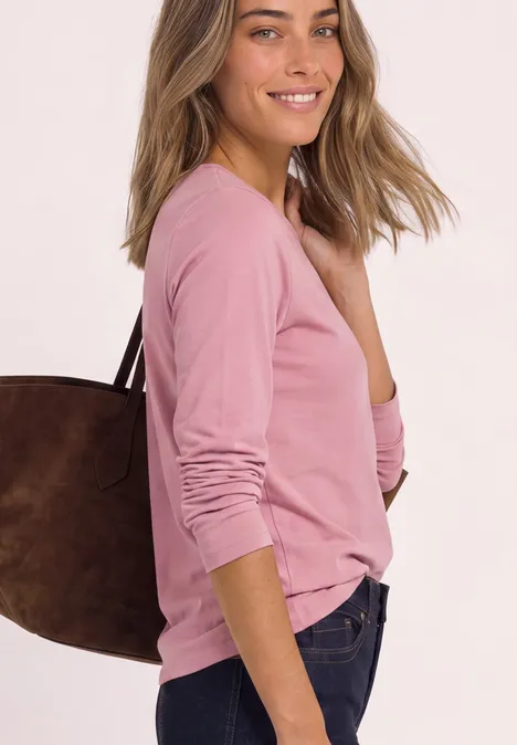 Frau in rosa Langarmshirt mit brauner Schultertasche lächelt seitlich in die Kamera.