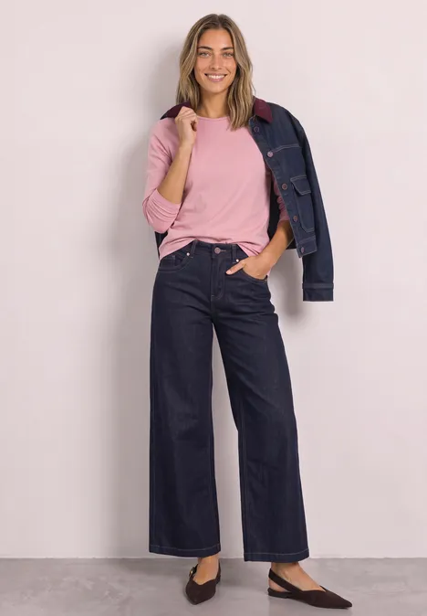 Frau in rosafarbenem Shirt, lässiger Jeansjacke über der Schulter, weiten, dunklen Jeans und Ballerinas.