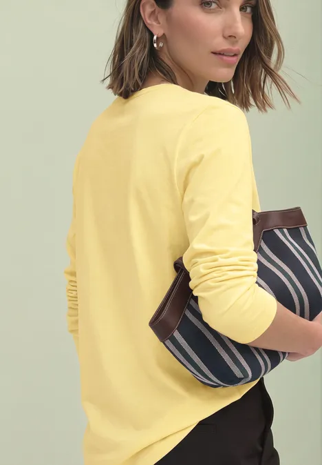 Eine Frau im gelben Langarmshirt hält eine blau-weiß gestreifte Tasche und blickt über die Schulter.