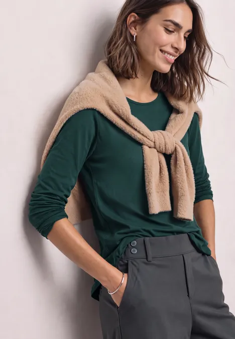 Basic Langarm Shirt in Unifarbe shadow teal Frau in grünem Langarmshirt mit beiger Strickjacke um die Schultern und grauer Hose, lächelnd.
