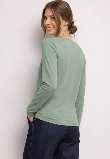 T-shirt basique à manches longues, couleur unie eucalyptus green Une femme souriante en haut vert à manches longues et jean regarde par-dessus son épaule.