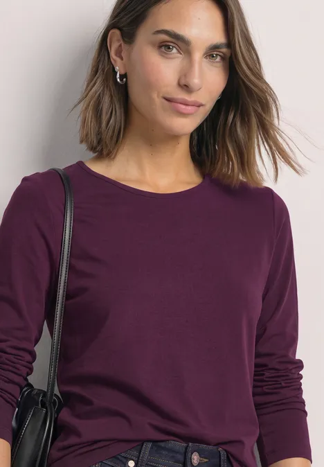 T-shirt basique uni à manches longues berry maroon Junge Frau trägt ein bordeauxfarbenes Langarmshirt und eine schwarze Schultertasche, vor hellem Hintergrund.