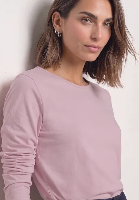 T-shirt basique uni à manches longues quiet rose Frau lehnt lässig an Wand, trägt rosafarbenes Langarmshirt und silberne Creolen.