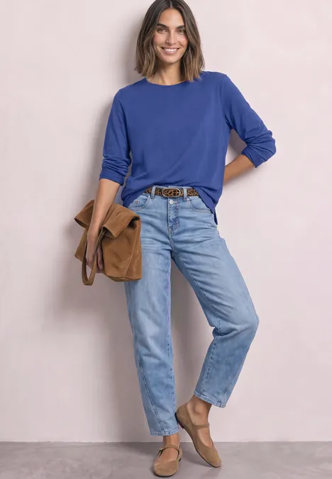 Frau in blauem Pullover und Jeans mit brauner Tasche und Schuhen vor neutralem Hintergrund.