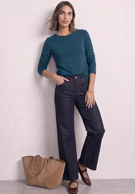 Frau in tealfarbenem Shirt, schwarzen Jeans und braunen Loafers, daneben eine große Tasche vor hellem Hintergrund.