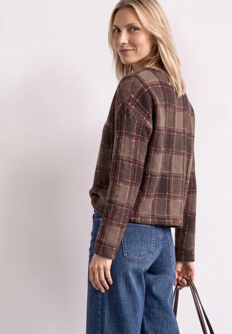 Eine Frau trägt eine braune Karojacke, blaue Jeans und eine Handtasche, schaut über die Schulter.