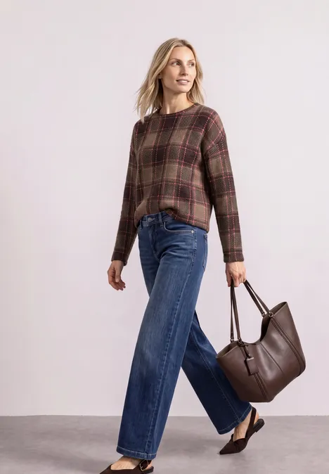 Frau mit karierter Bluse, weiten Jeans und brauner Ledertasche vor hellem Hintergrund.