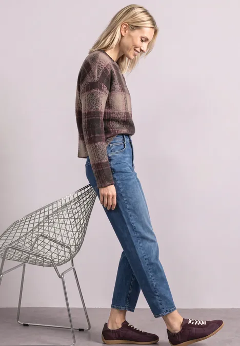 Frau mit kariertem Pullover und Jeans lehnt entspannt an einem Drahtstuhl.