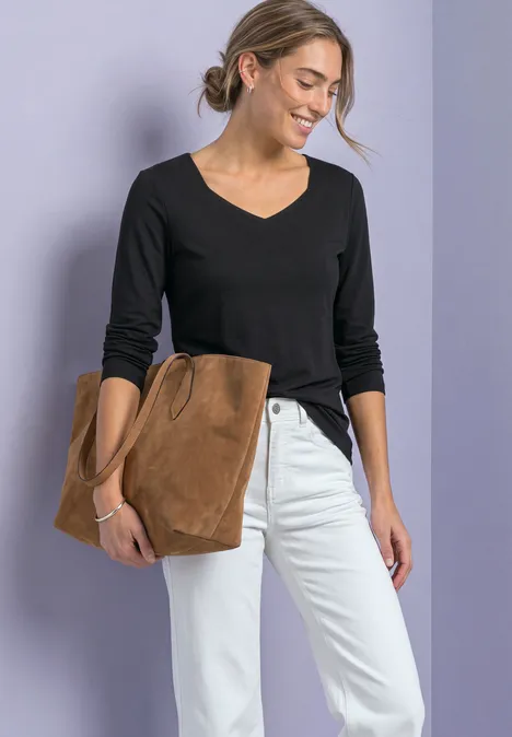 Femme souriante en pull noir à col V, jean blanc, tenant une grande pochette en daim marron.