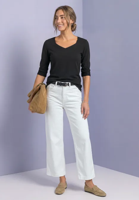 Femme en top noir, jean blanc, ceinture noire, sac marron et mocassins beiges sur fond neutre.