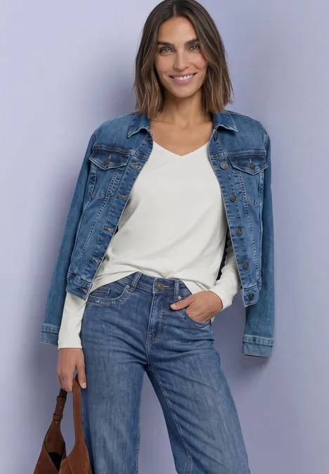 Une femme souriante pose devant un fond clair, veste en jean sur les épaules, t-shirt blanc et jeans bleu