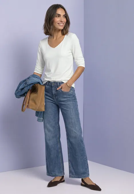 Femme en tee-shirt blanc à manches 3/4 et jean large, tenant deux vestes, sur fond violet.