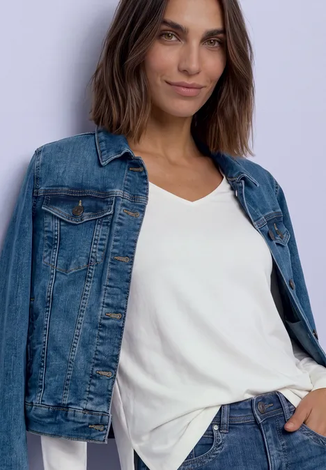 Femme portant une veste en jean et un t-shirt blanc, posant décontractée devant un fond clair.