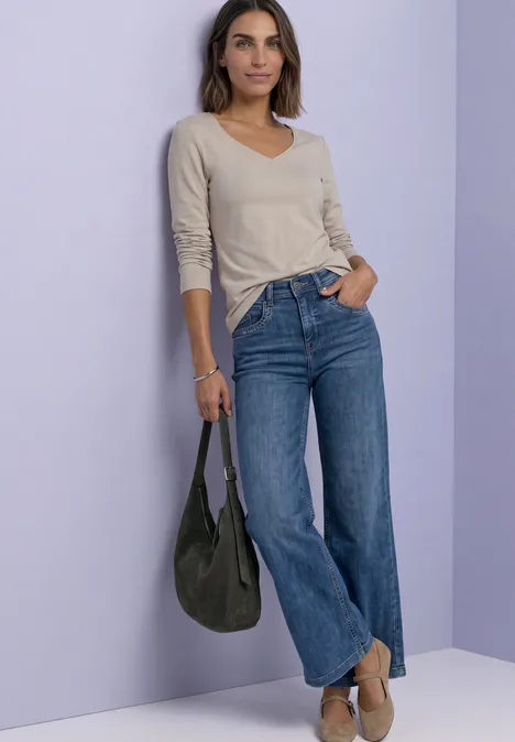 Femme appuyée sur un mur violet, porte un haut clair à manches longues, un jean ample et un sac sombre.
